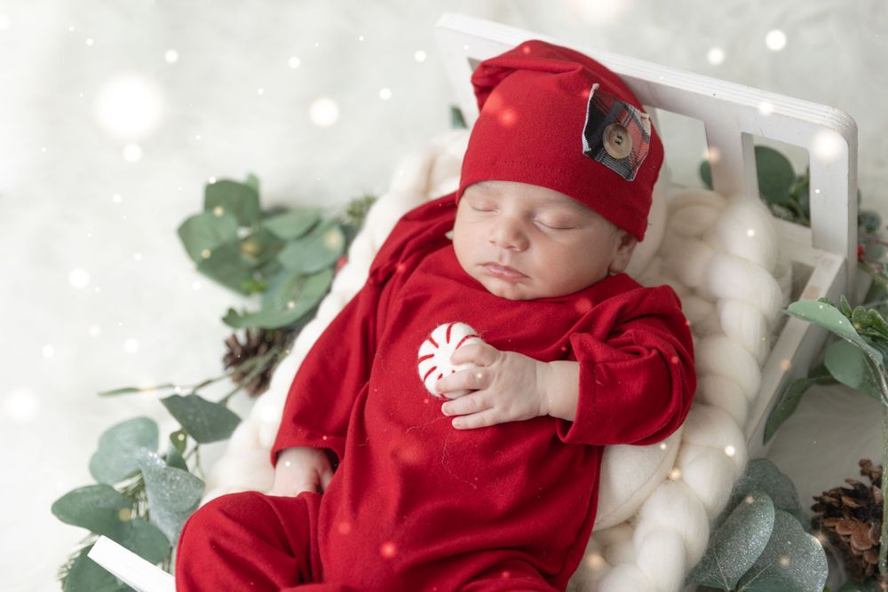 Sleeping newborn in bright red holiday outfit and hat lying on white backdrop with greenery.