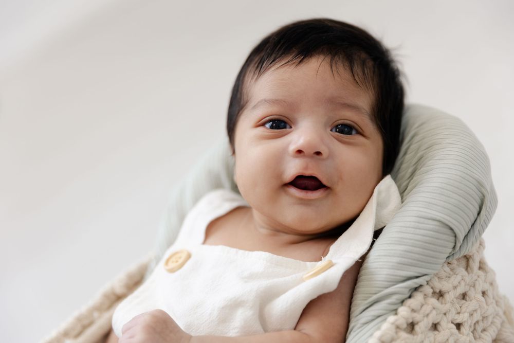 Baby wearing white outfit smiles while resting on cushioned support.