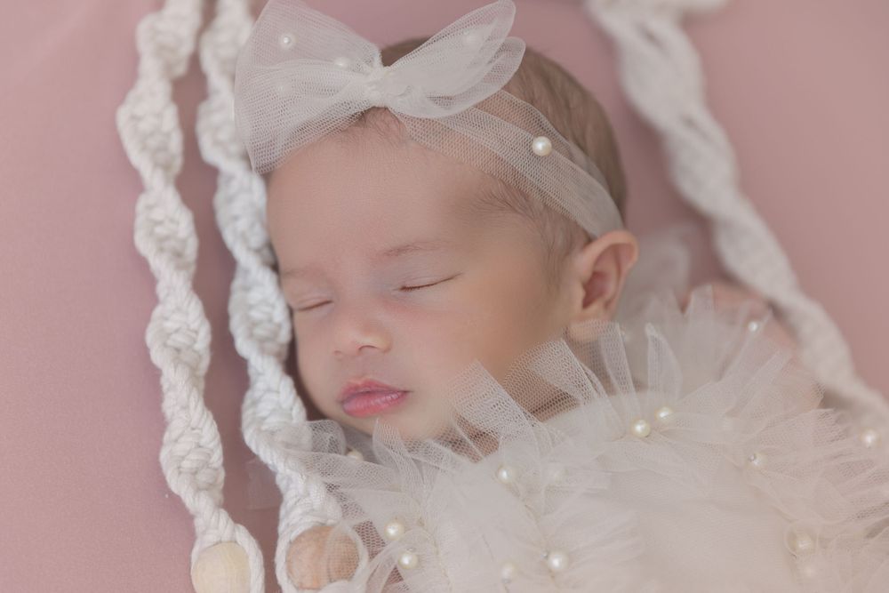 Peaceful baby in white lace outfit sleeping on macrame swing with pink background.