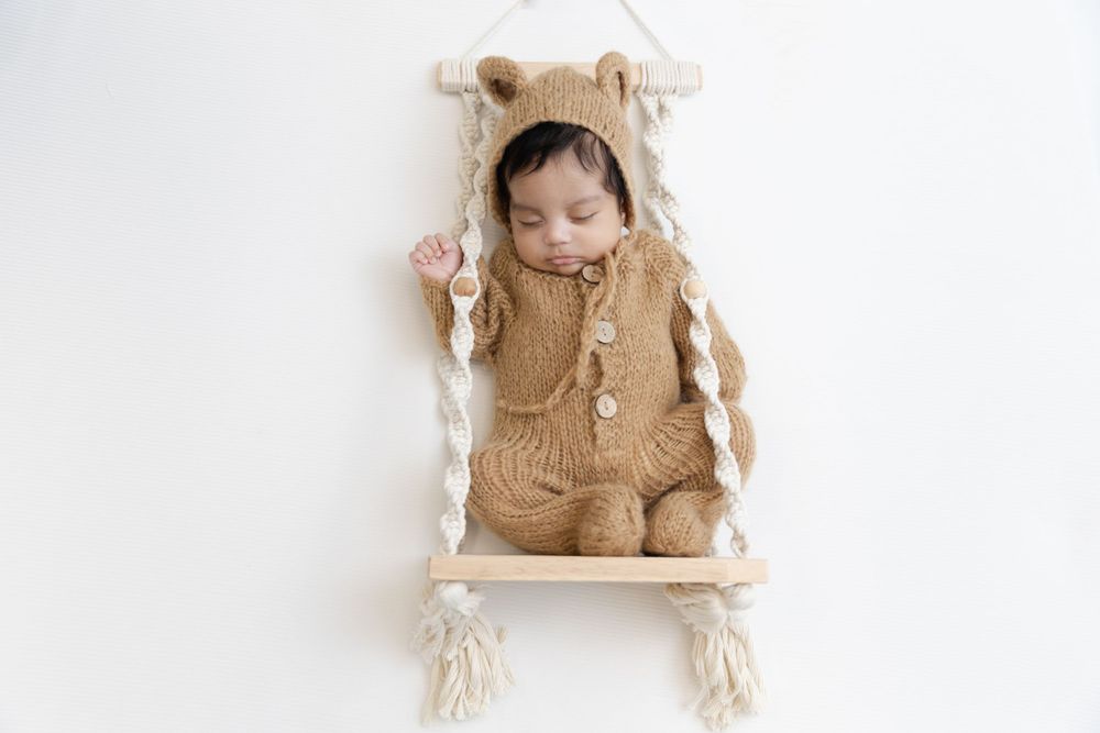Baby sleeping in cozy bear outfit on macrame swing against white wall.