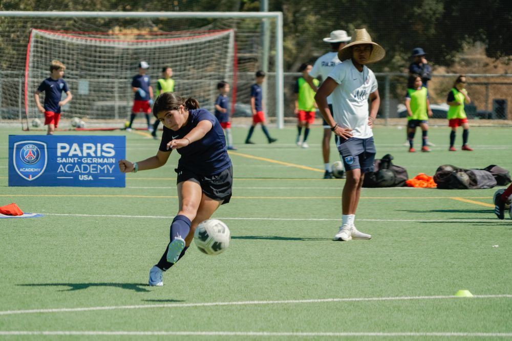Soccer player dribbling ball during practice session on outdoor turf field with goals visible.