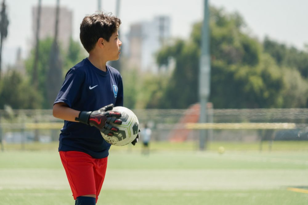 Young athlete in navy and red uniform holding soccer ball on sunny training field.