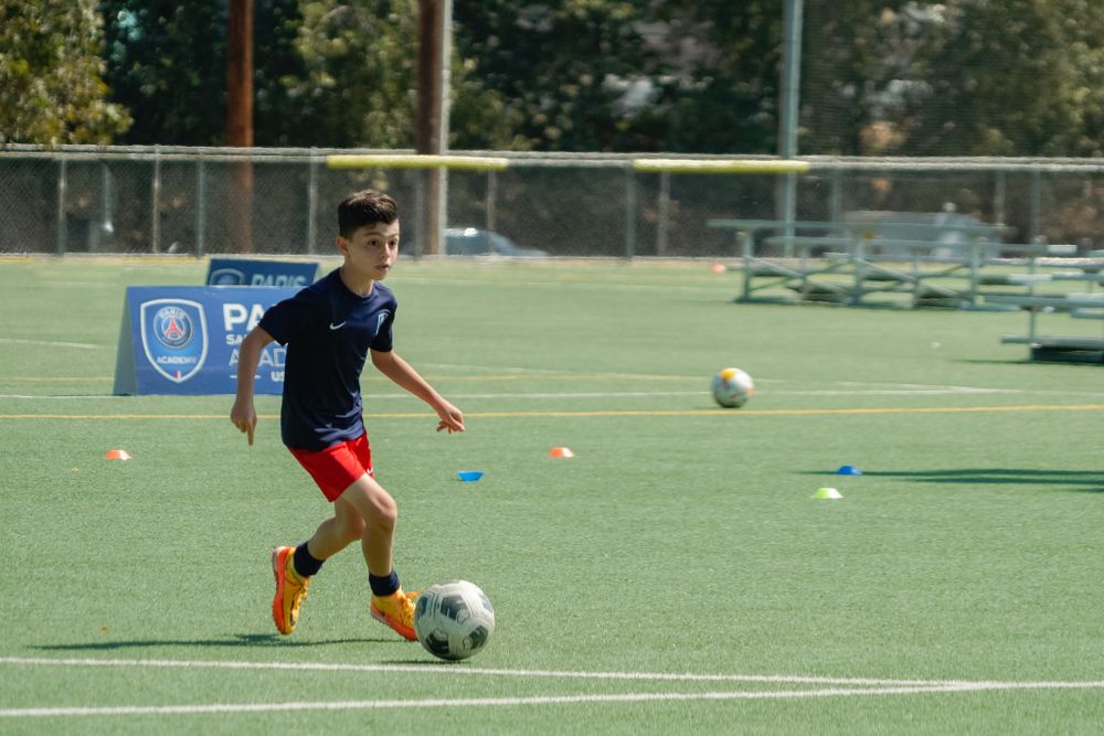 Youth soccer player in navy jersey and red shorts practicing with ball on artificial turf field.