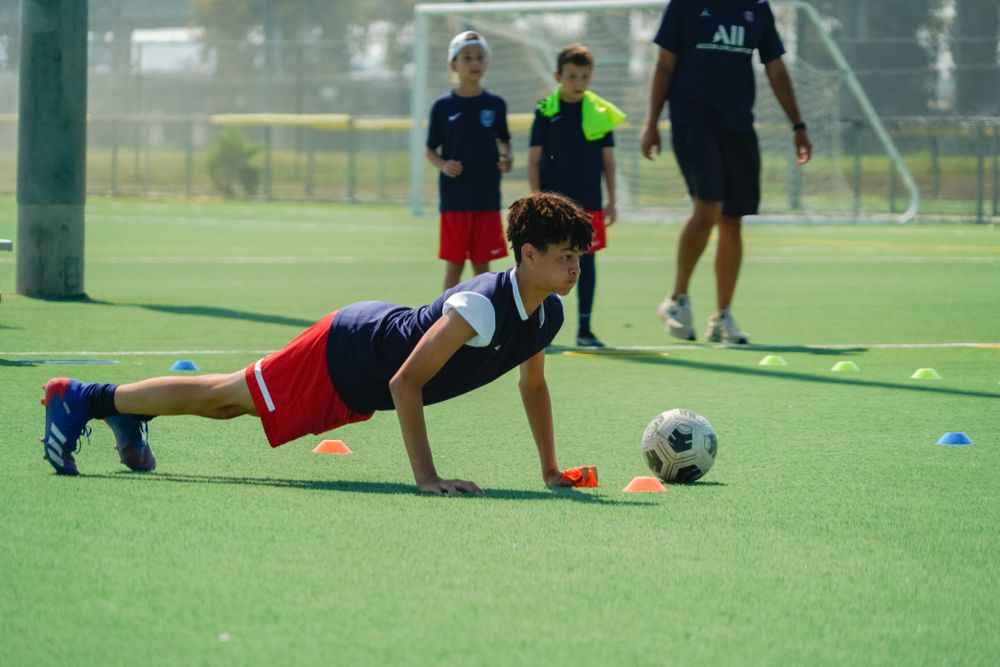 Young soccer player performing pushup drill with ball on artificial turf field.