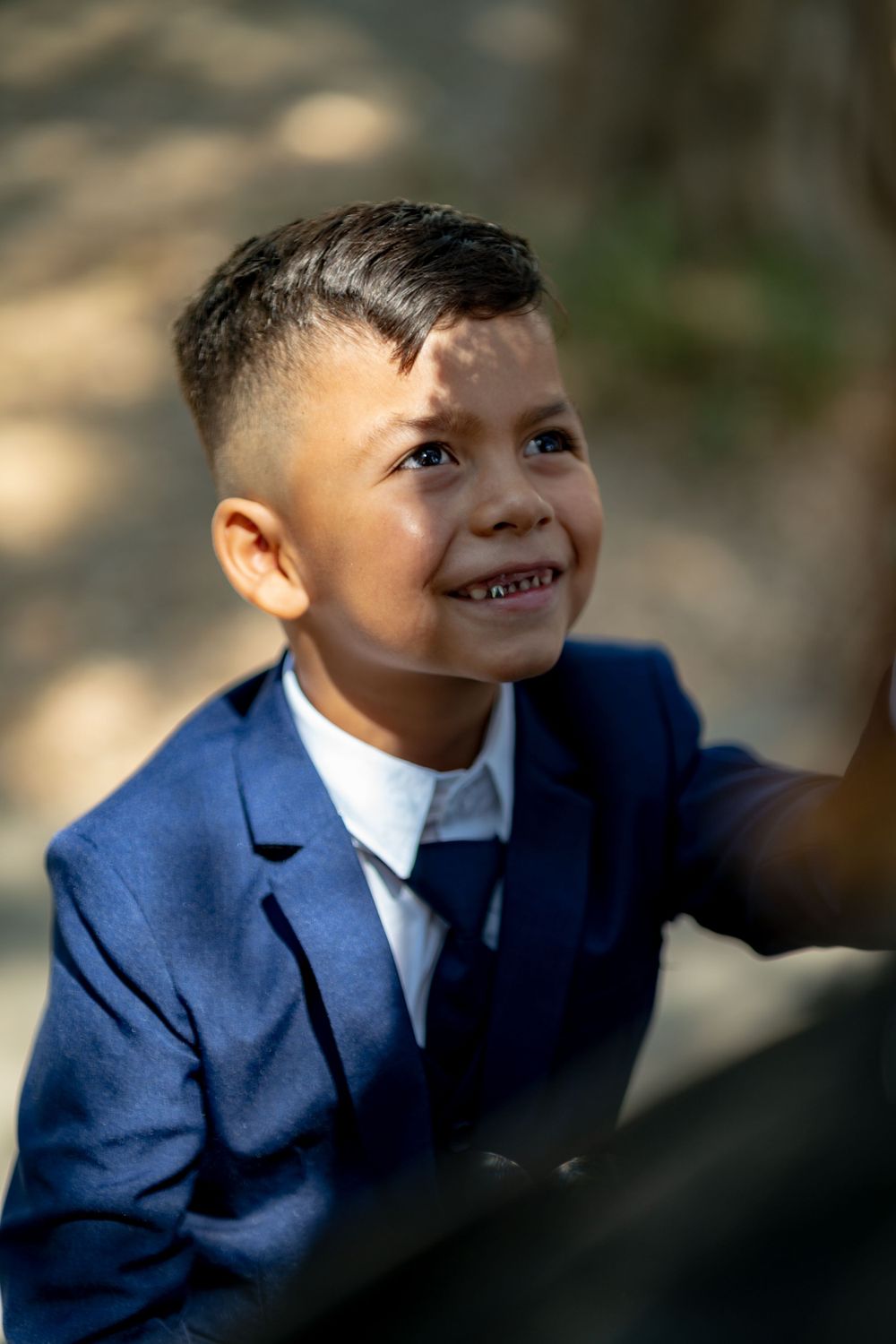 Young boy in navy blue suit smiling brightly in natural outdoor lighting.