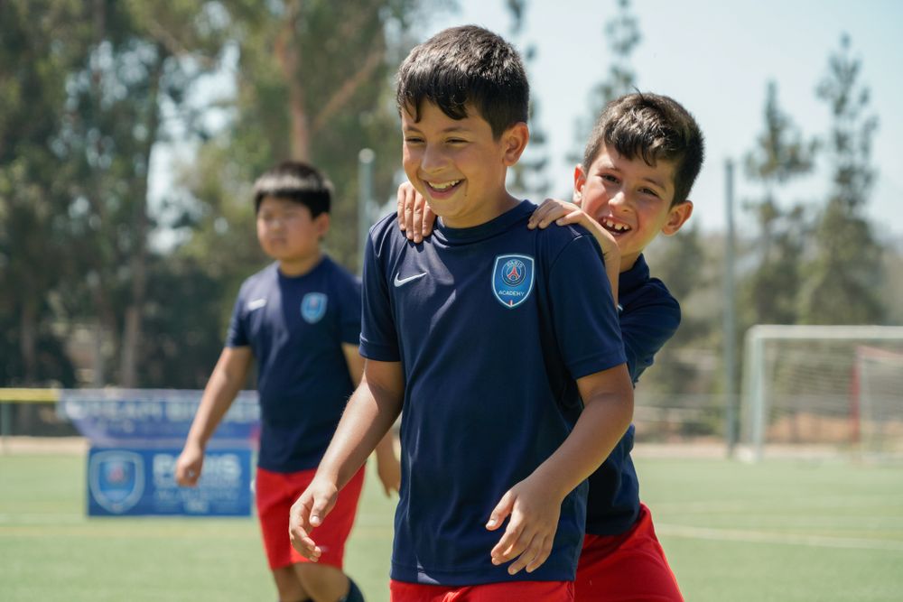 Young soccer players in navy uniforms share a lighthearted moment during practice on a sunny field.