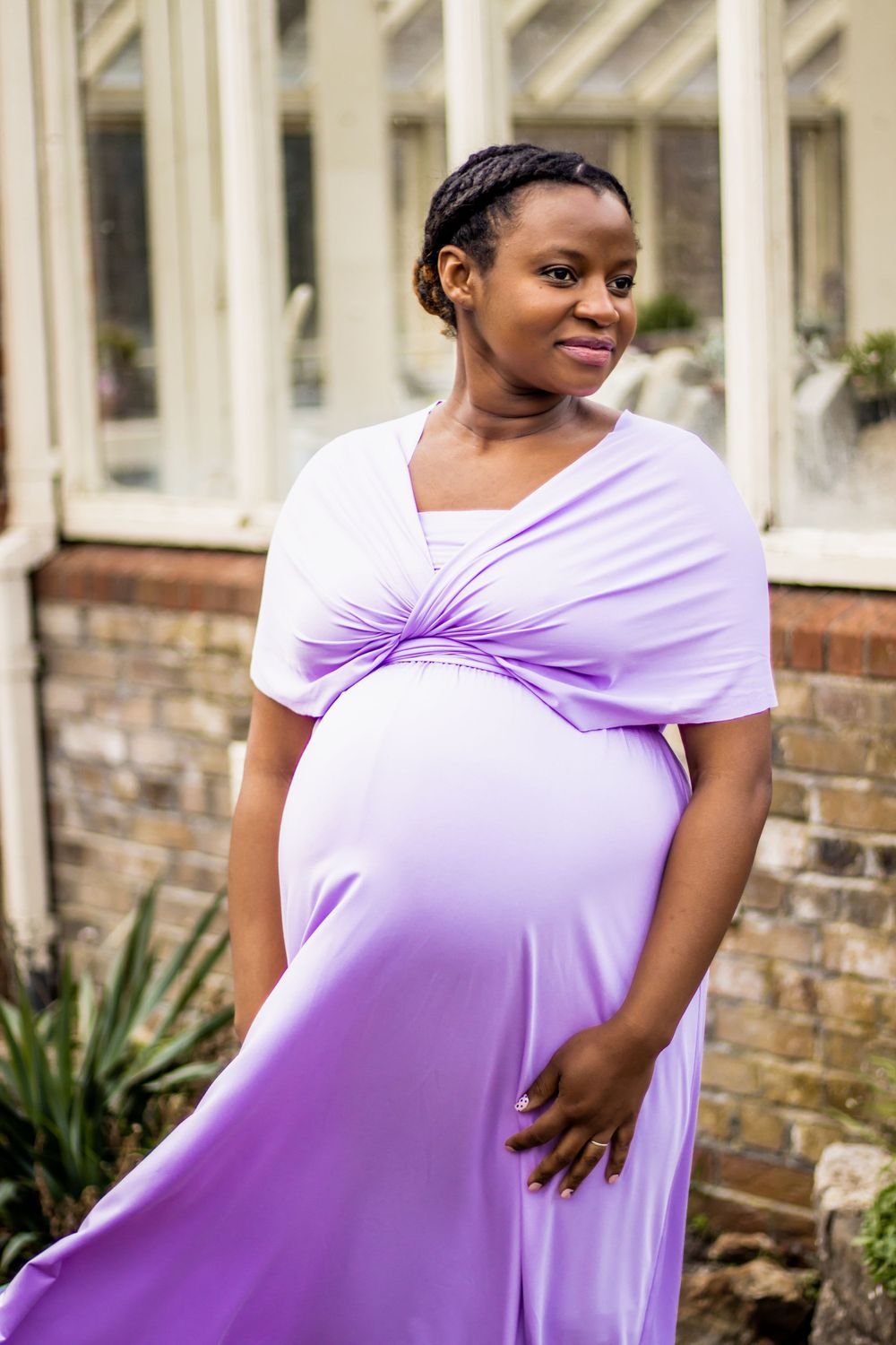 A glowing maternity photo featuring a flowing lilac dress against a backdrop of white columns and brick walls.