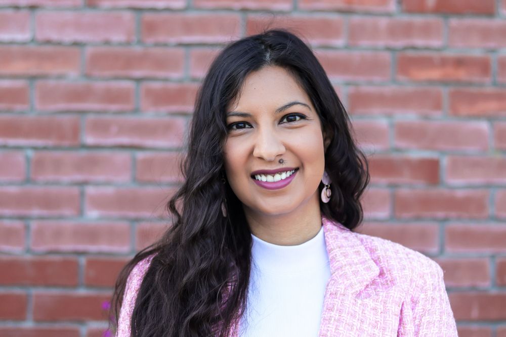 Cheerful closeup portrait with long dark hair wearing a pink blazer against brick wall.