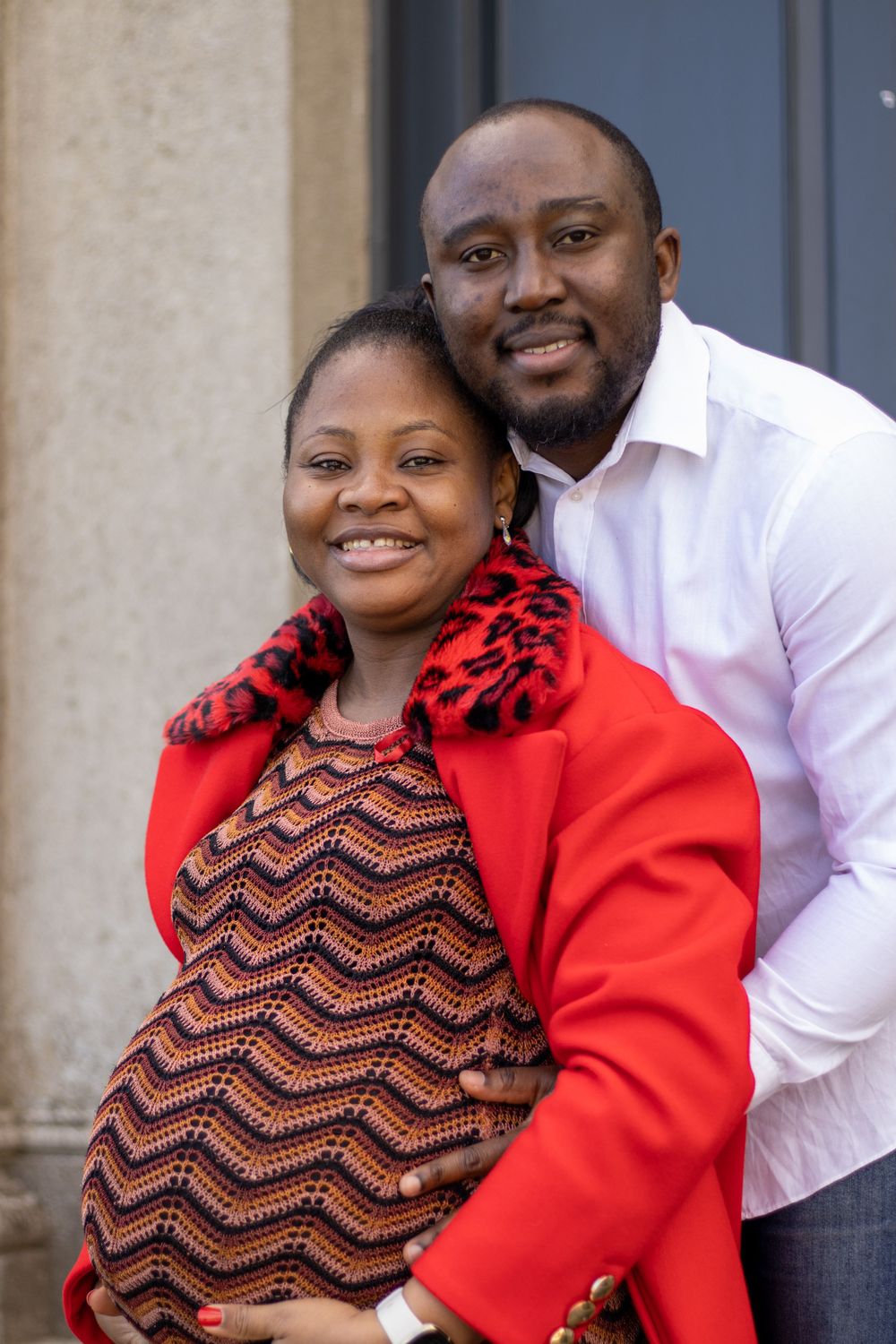 A loving pregnancy portrait of a couple, with a vibrant red coat and chevron pattern dress.