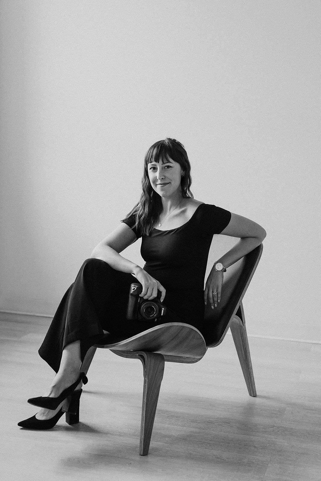 A photographer poses elegantly in a modern chair while holding a camera.