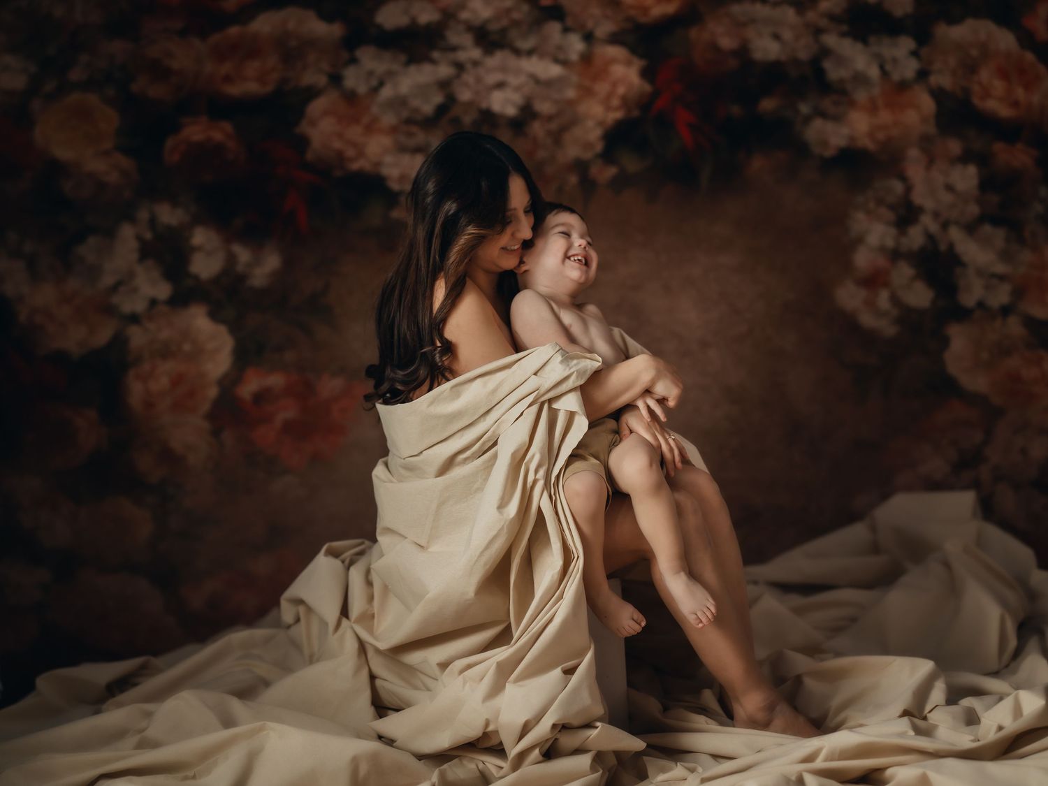 Mother and baby wrapped in flowing fabric, sitting in an elegant floral studio setting. Toronto motherhood photography.