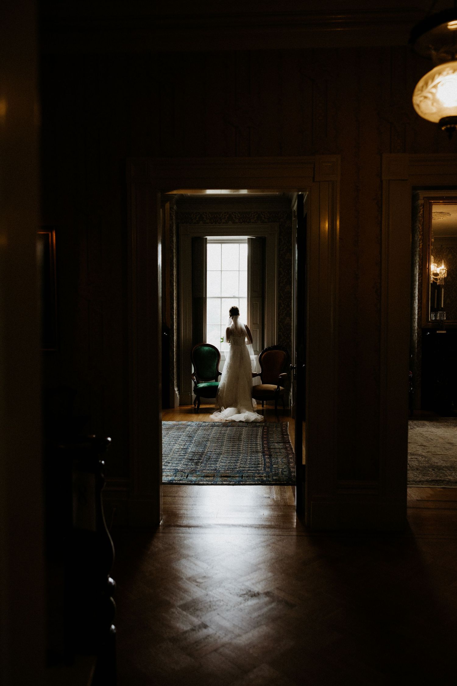 Bride in white gown stands silhouetted by window in elegant dark hallway of historic mansion. Rotch Jones Duff House Miranda Photo & Co