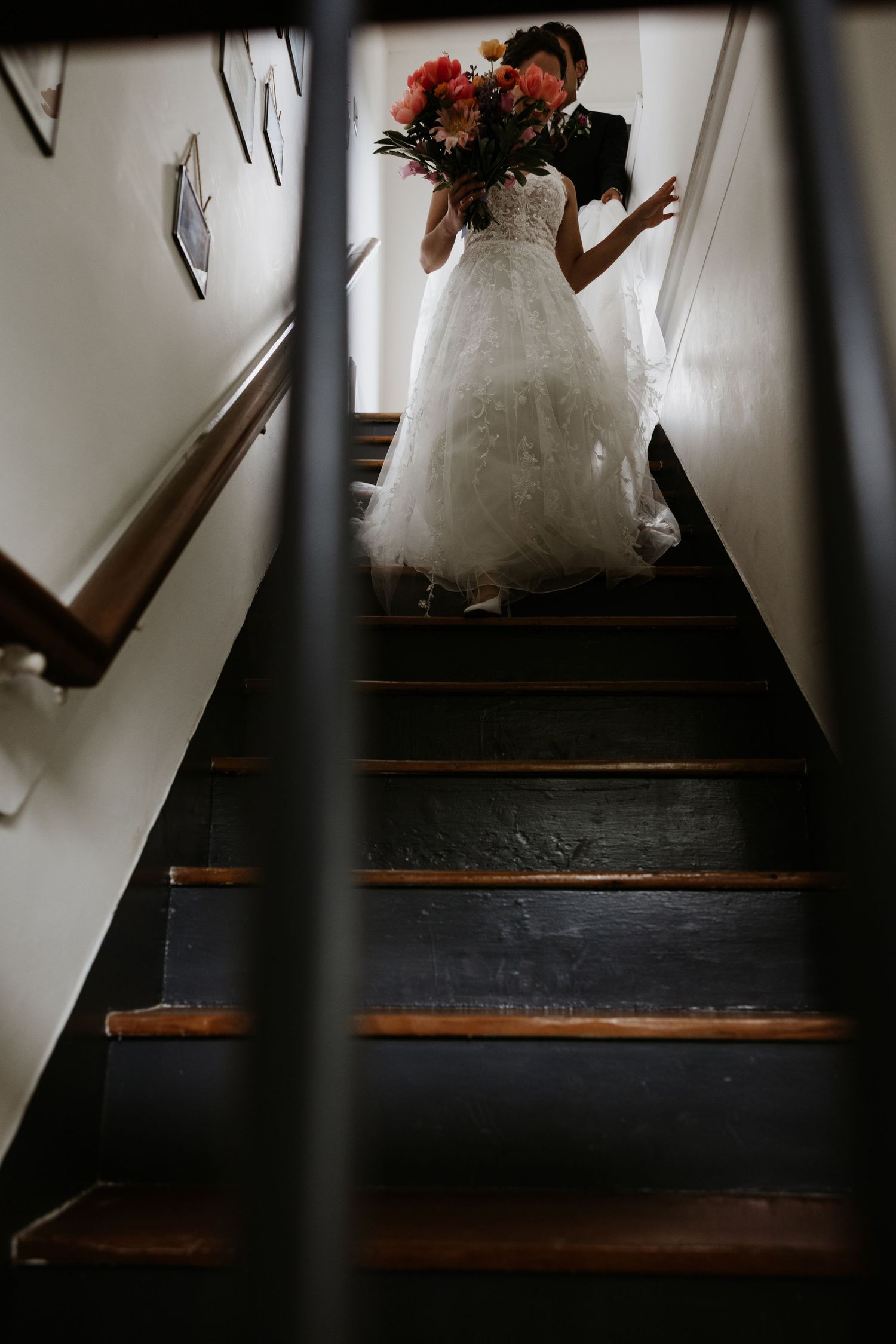 Bride in white lace gown holding colorful bouquet stands on dark wooden staircase with groom behind her. New Bedford, Ma. Miranda Photo & Co