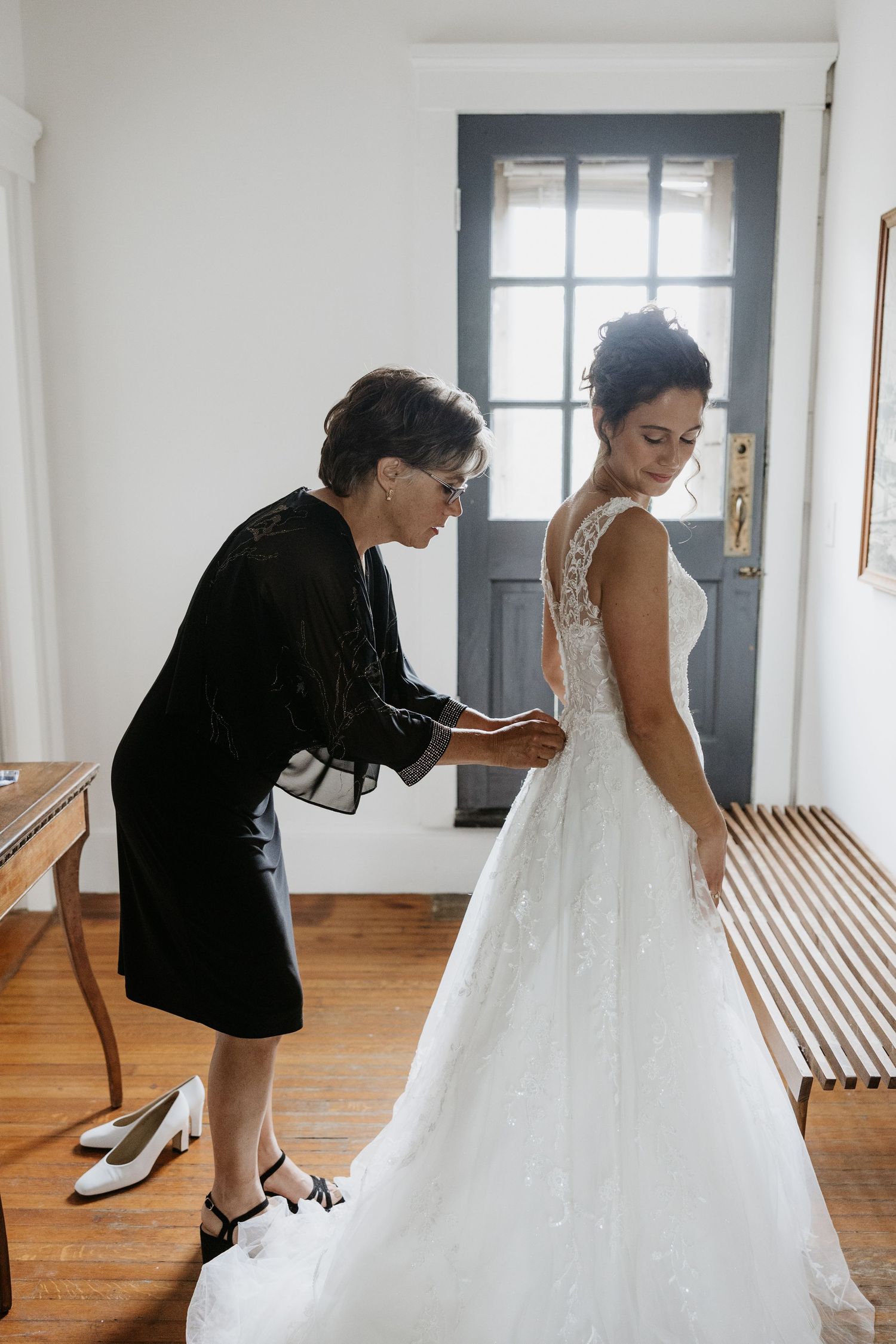 Mother helping bride button up her lace wedding dress in a bright white dressing room. Rotch Jones Duff House Miranda Photo & Co