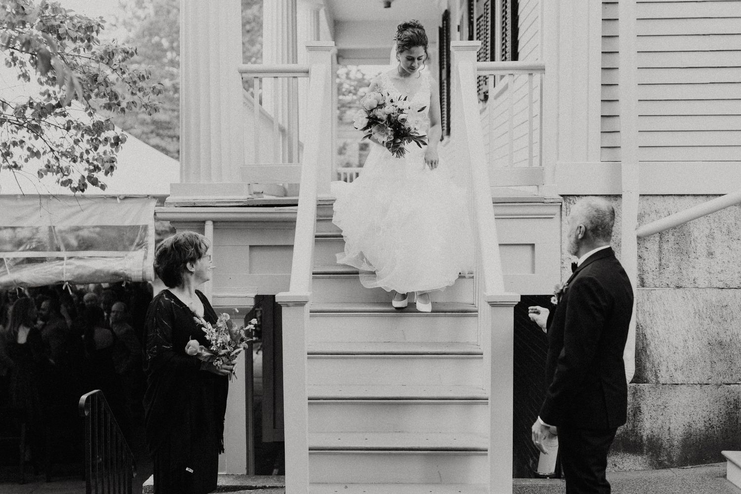 Bride in flowing white gown descends porch stairs holding bouquet with family waiting below. New Bedford, Ma. Miranda Photo & Co