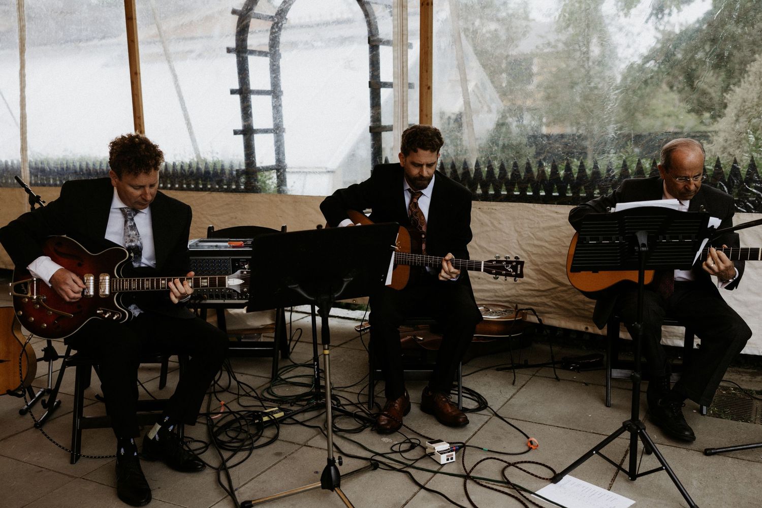 Three musicians playing acoustic guitars at an outdoor wedding reception under a tent. Rotch Jones Duff House Miranda Photo & Co