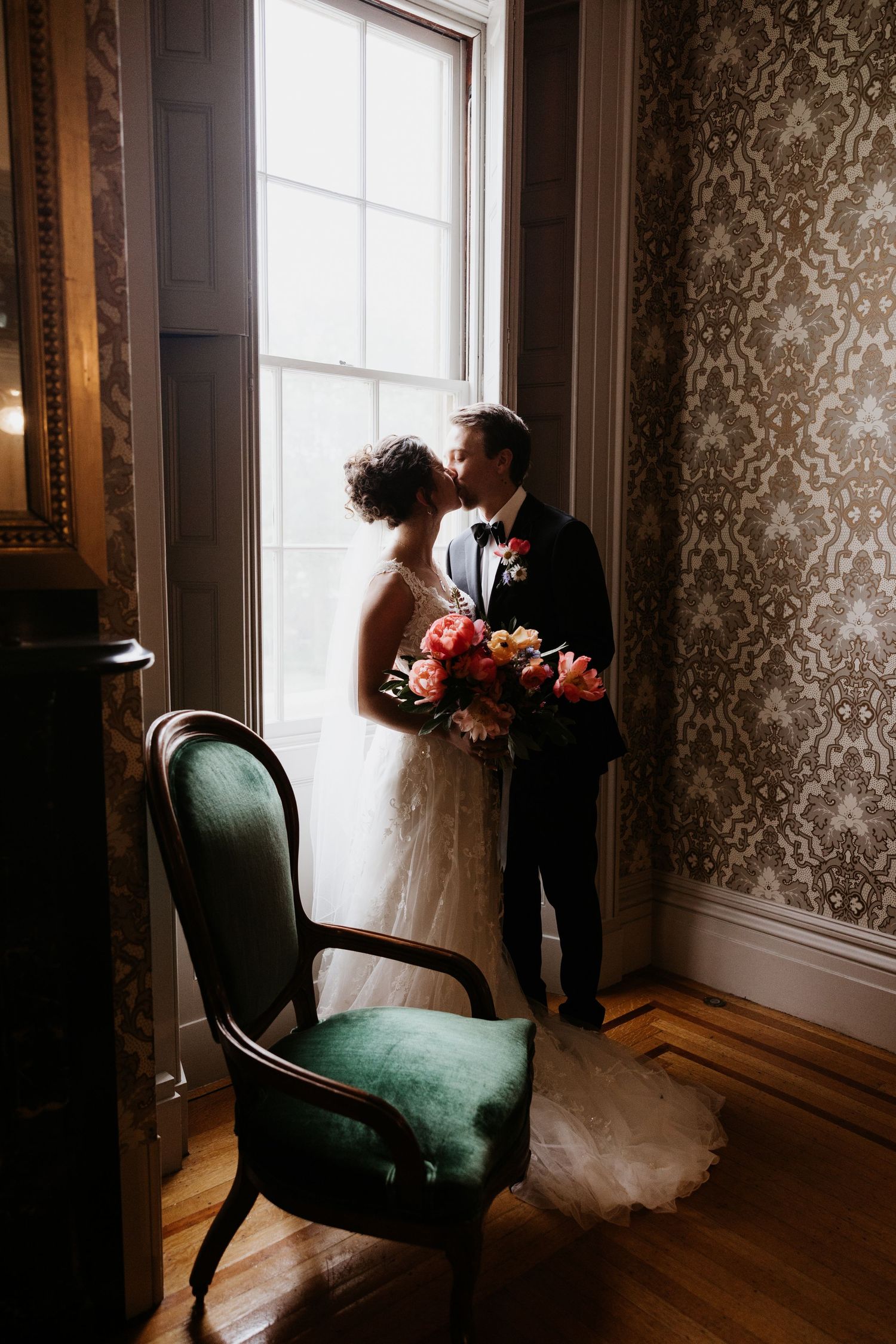 Bride and groom sharing a romantic kiss by a window in an elegant wallpapered room on their wedding day. New Bedford, Ma. Miranda Photo & Co