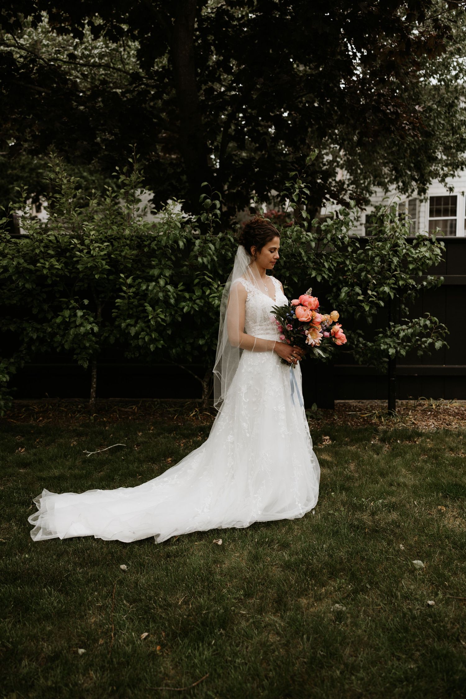 Bride in a white lace gown with veil holding a colorful bouquet in a lush garden setting. New Bedford, Ma. Miranda Photo & Co