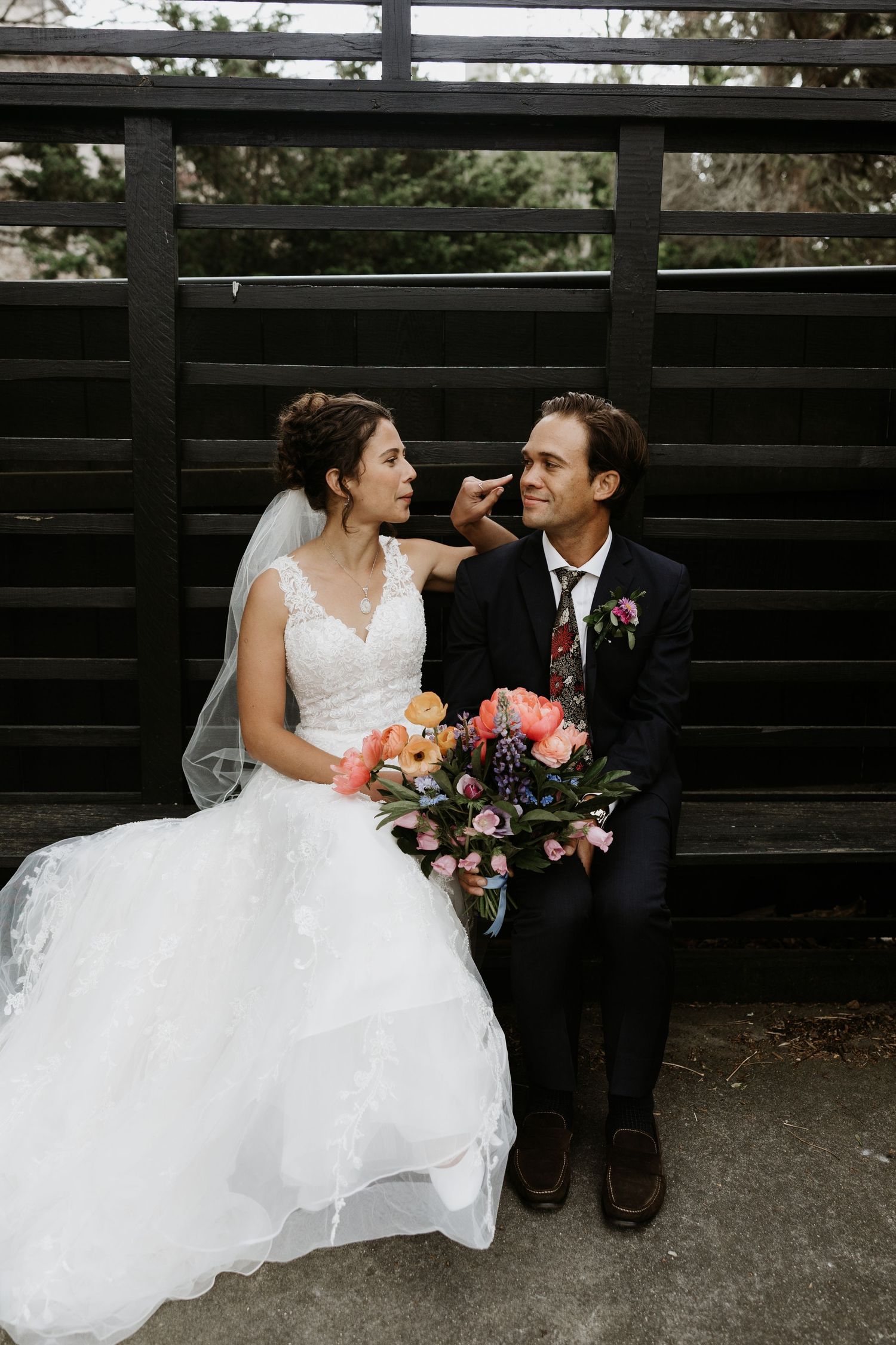 Bride and groom sitting together on a bench, gazing at each other, holding a colorful bouquet of pink and orange flowers. New Bedford, Ma. M