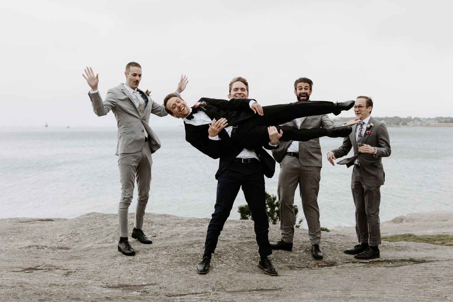 Groomsmen in gray suits playfully lift groom on rocky New England waterfront on wedding day. New Bedford, Ma. Miranda Photo & Co