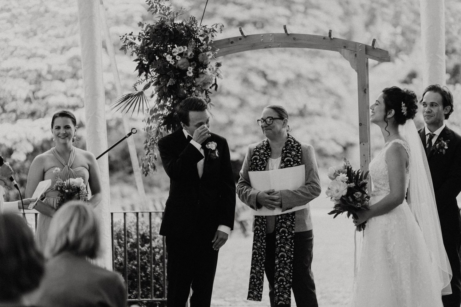 Black and white outdoor wedding ceremony with bride, groom, and officiant under a wooden arbor. New Bedford, Ma. Miranda Photo & Co
