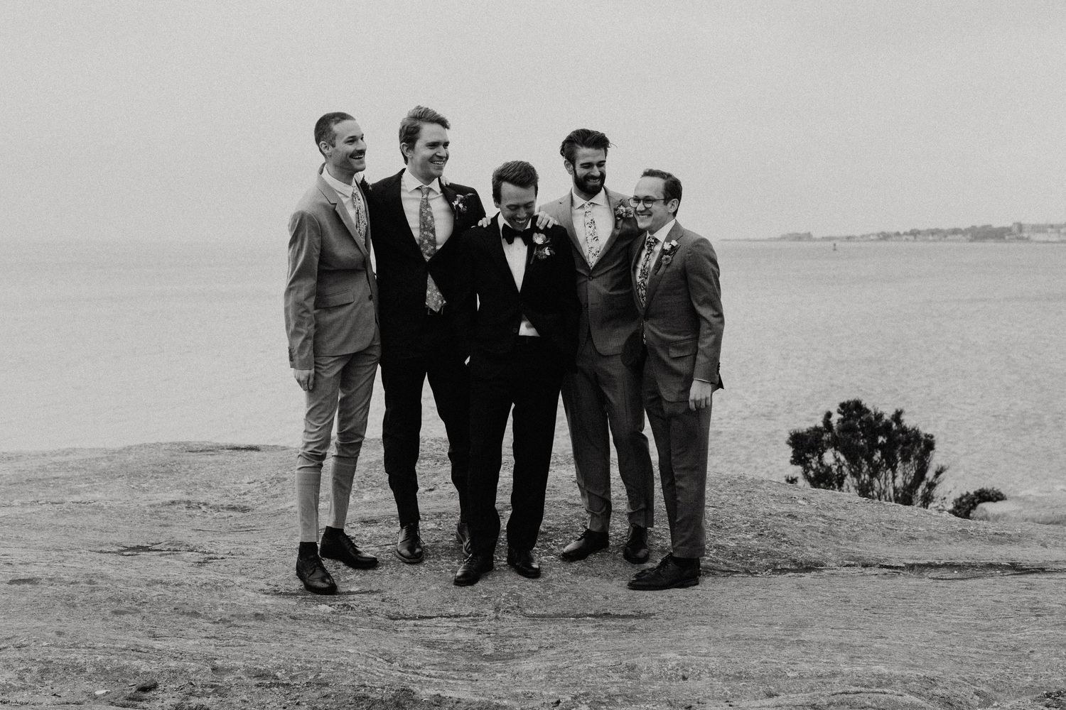 Black and white photo of groom and groomsmen laughing together outdoors near a waterfront. New Bedford, Ma. Miranda Photo & Co