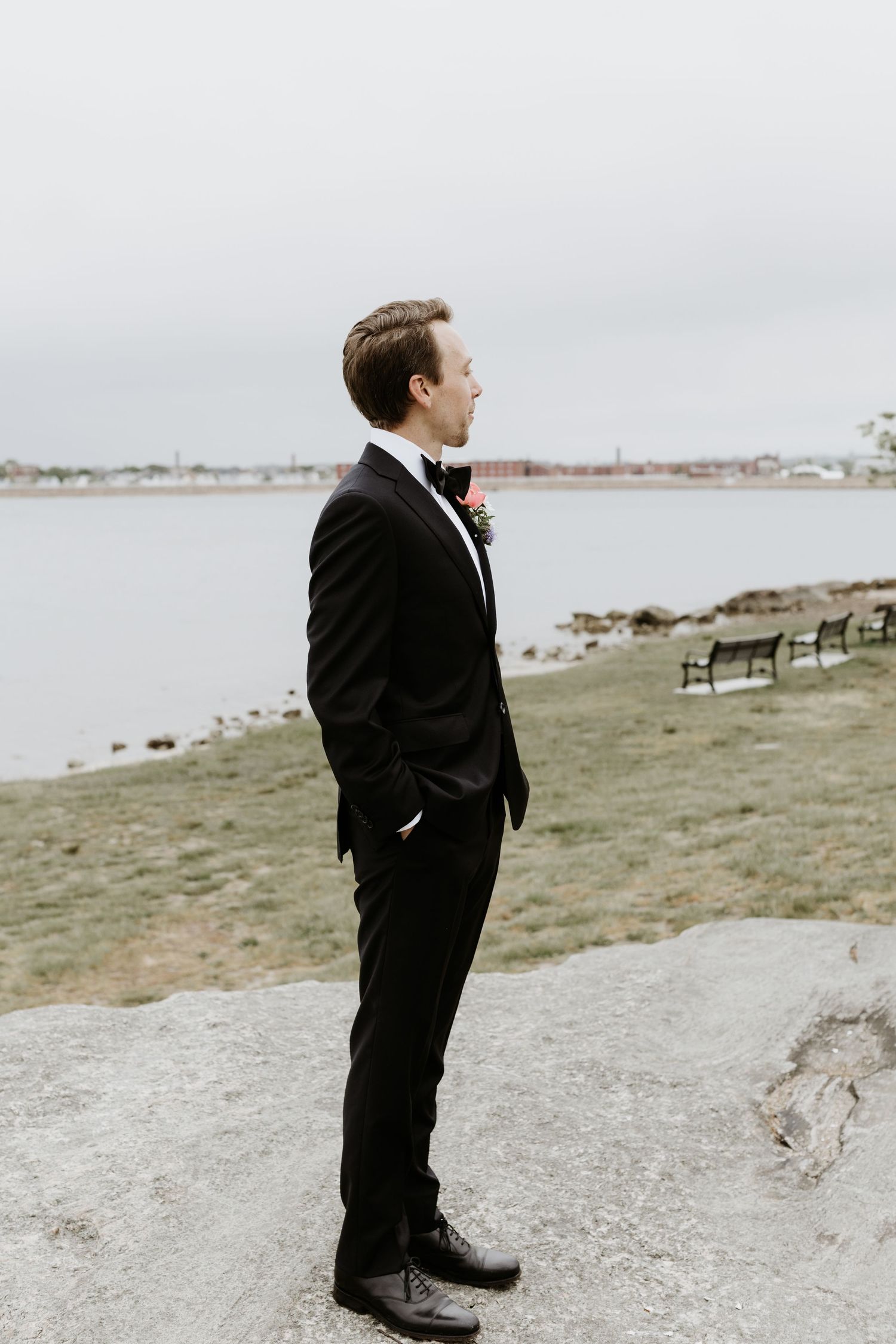 Dapper groom in black tuxedo standing by waterfront on his wedding day looking into the distance. New Bedford, Ma. Miranda Photo & Co