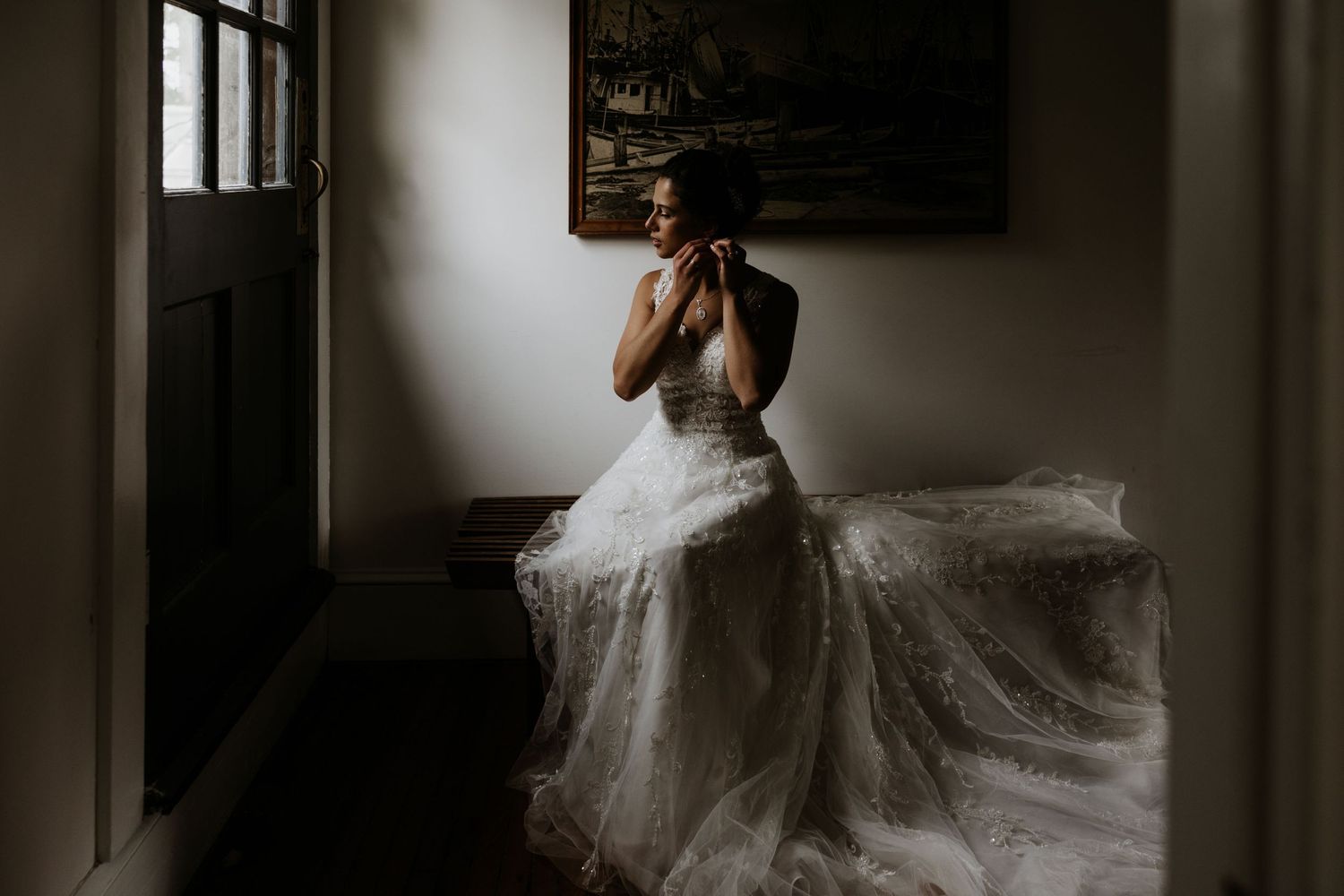 Bride in lace gown sits in moody light adjusting earrings beside antique door and painting. New Bedford, Ma. Miranda Photo & Co