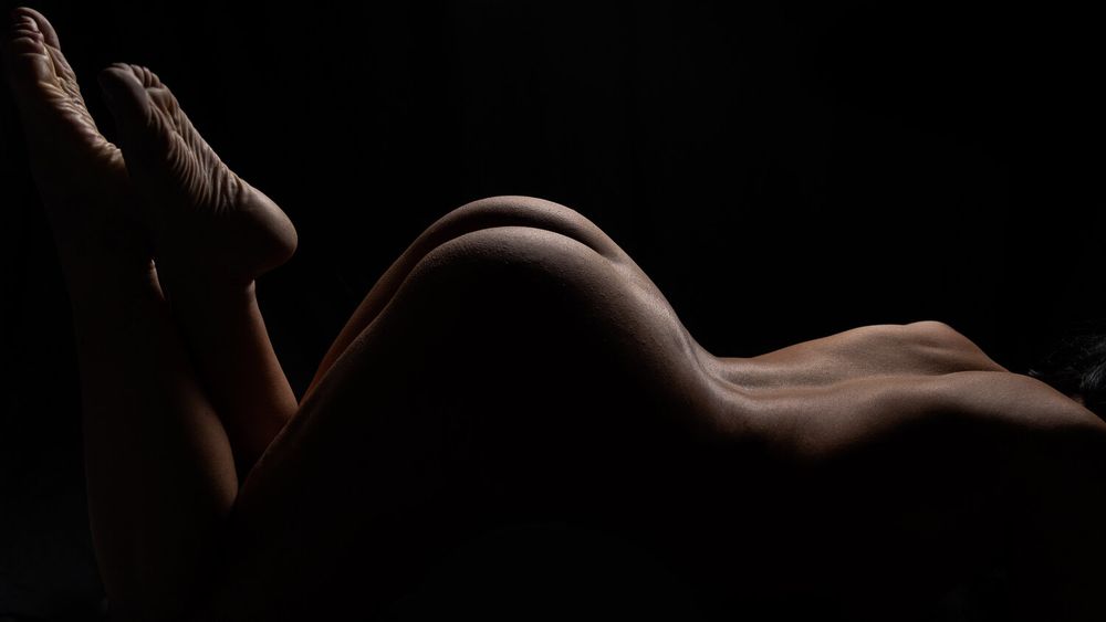 Artistic nude black and white photograph showing graceful curves and shadows against dark background.