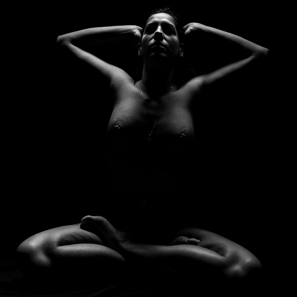 Low key nude art photo showing silhouette against dark background with arms raised.