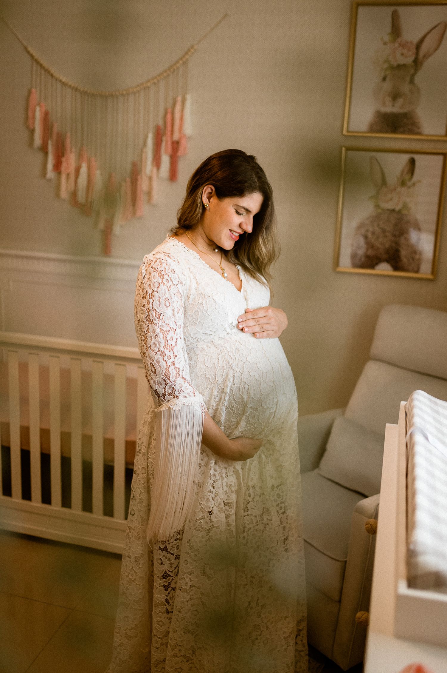 Expectant mother in white lace dress poses in nursery with soft decor.