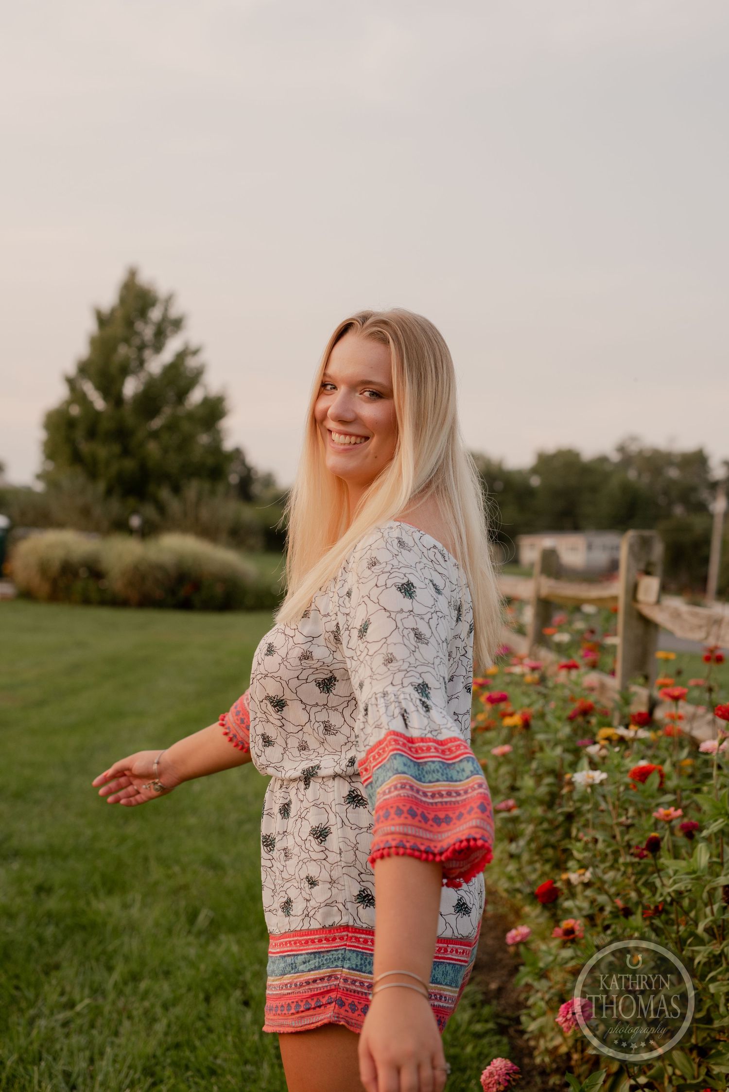 Woman in patterned romper with red trim stands by flowering garden during golden hour.