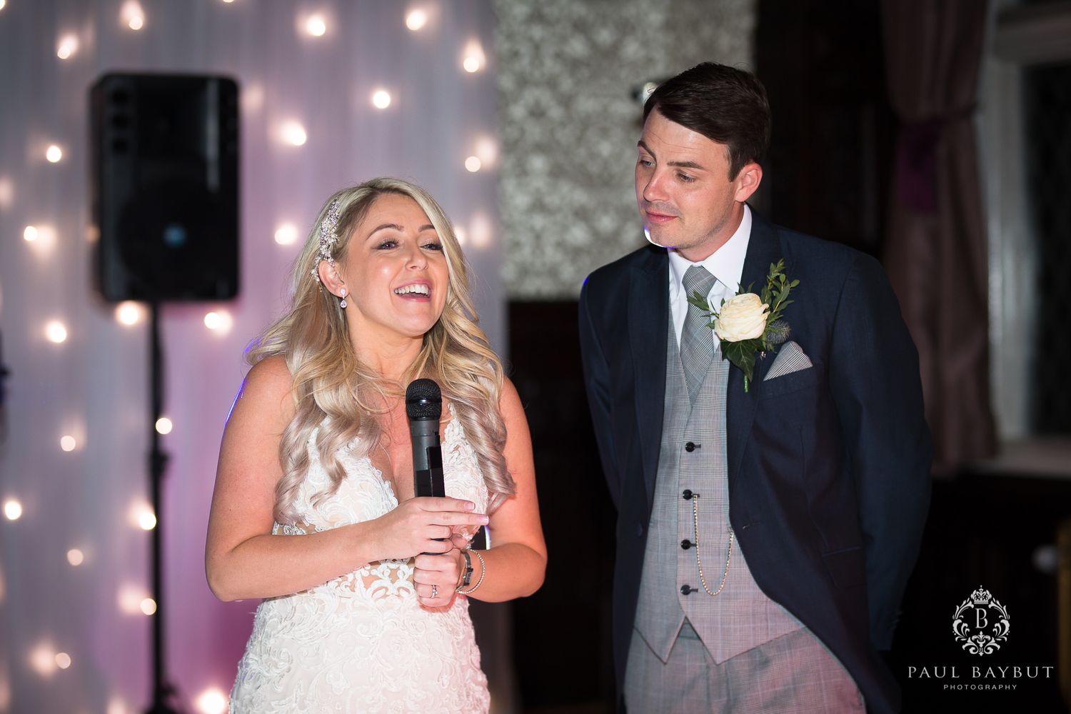 A couple shares a joyful moment during their wedding reception speech with twinkle lights.
