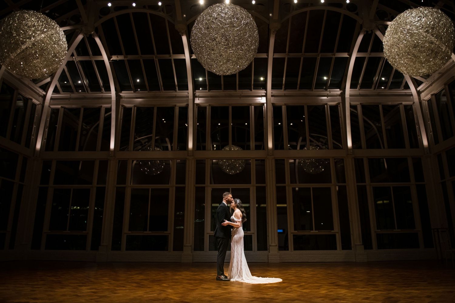 Bride and groom dance romantically under glowing sphere chandeliers in a grand ballroom.