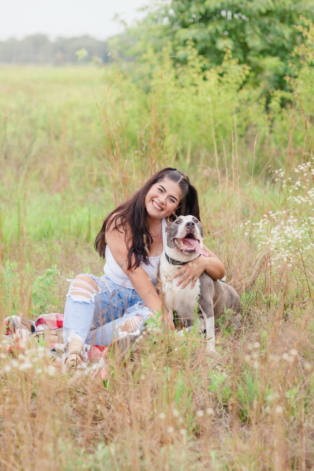 Woman in ripped jeans sits in a wildflower field with her happy pit bull dog.