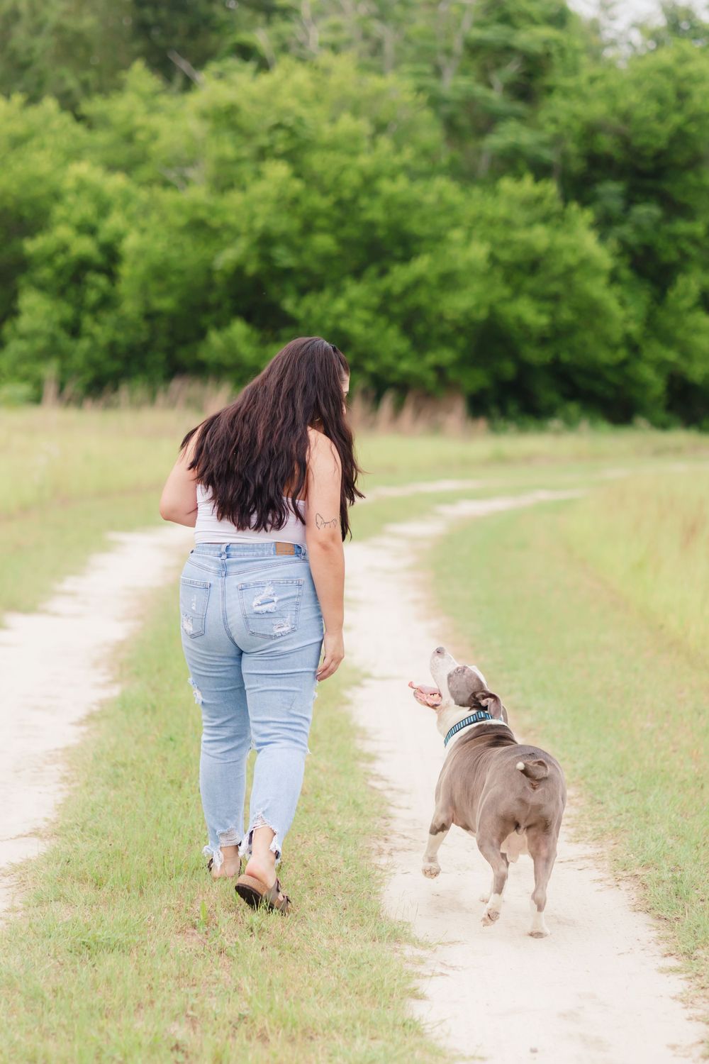 Woman in jeans walking down a dirt path with her dog following behind.