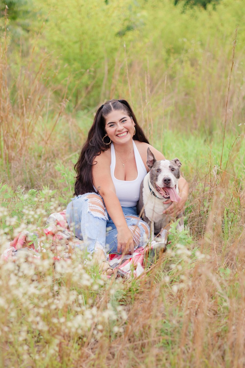 Woman in casual attire poses with her smiling pit bull dog in a sunny meadow.