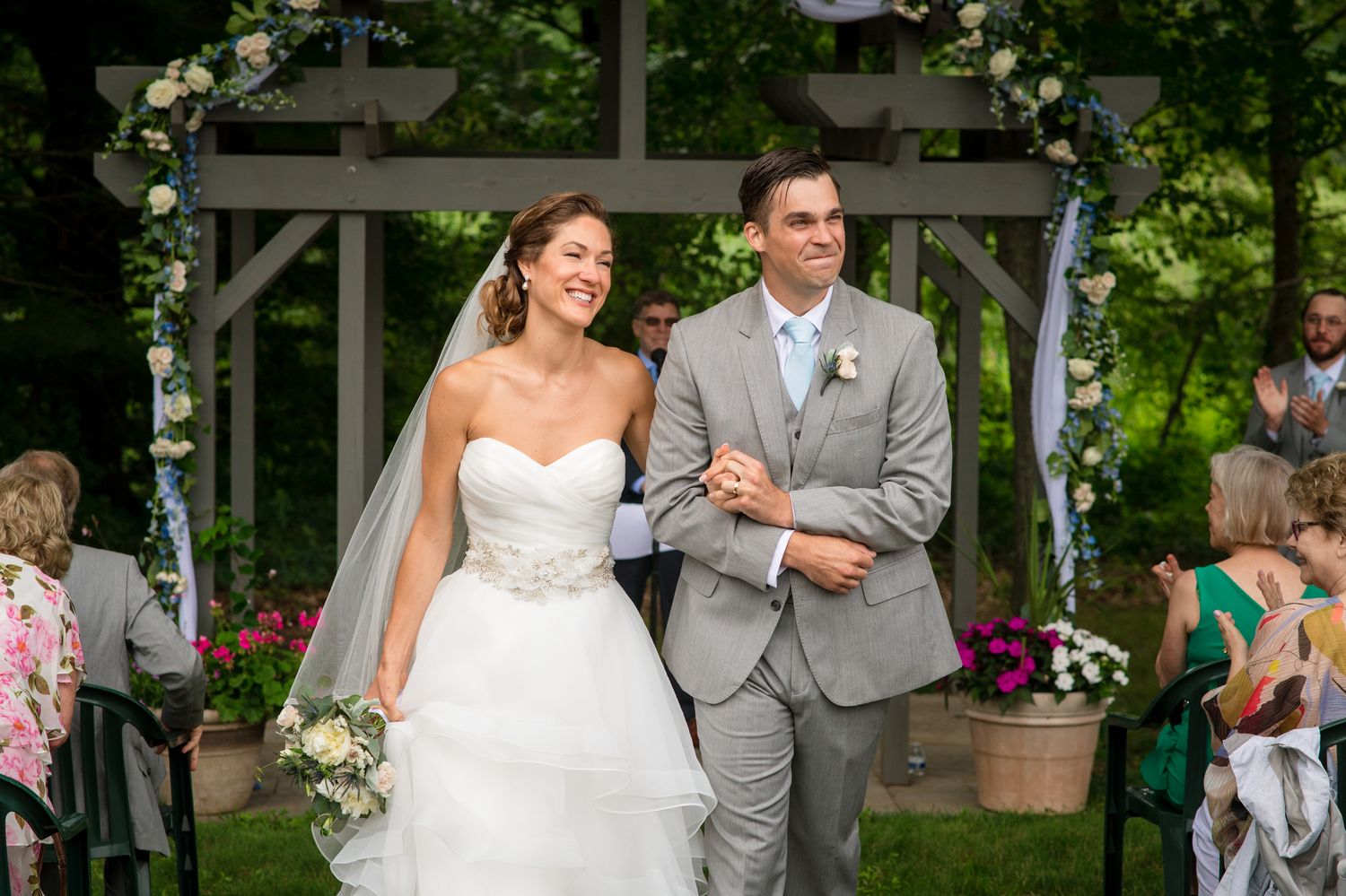 Outdoor wedding ceremony under a white pergola with lush greenery and floral decorations.