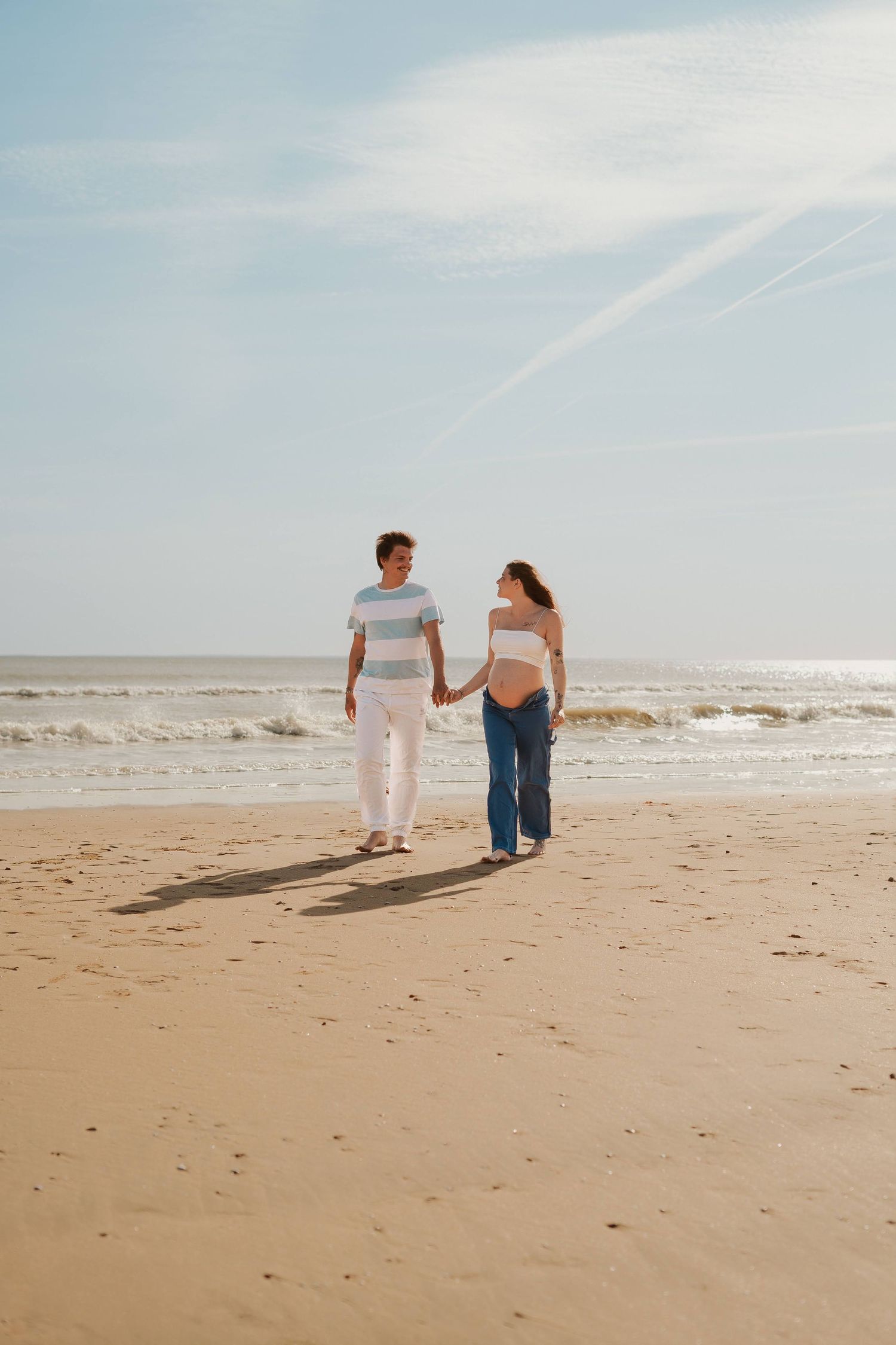 Couple walking hand in hand along sandy beach under blue sky.