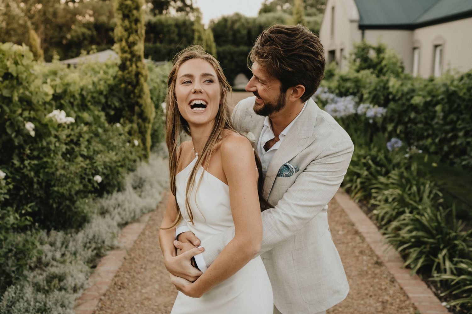 Candid moment of a couple laughing together in a garden pathway wearing wedding attire.
