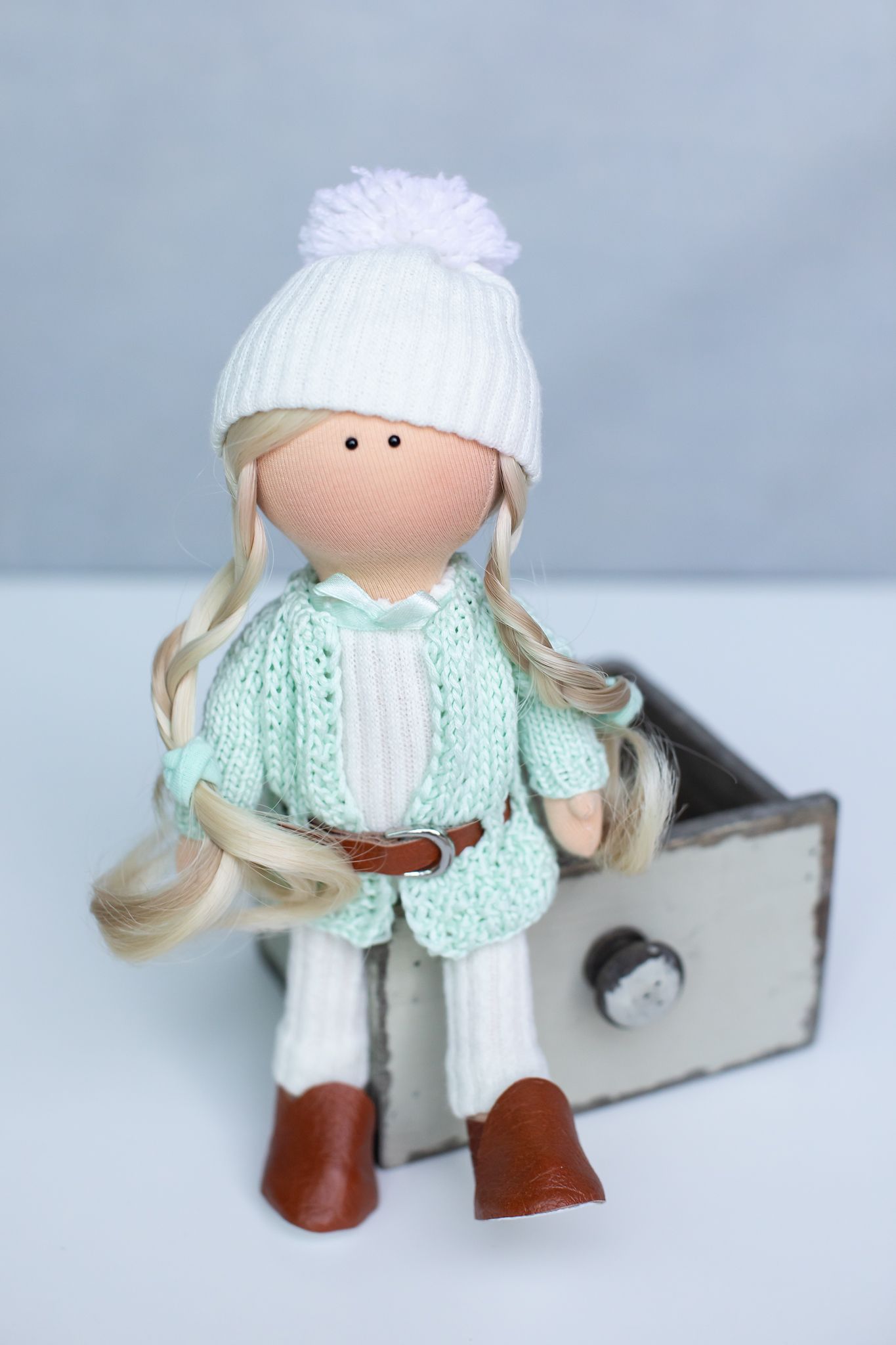 Handmade fabric doll with blonde braids in mint knit cardigan and white pom-pom hat sitting on a drawer.