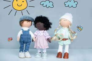 Three diverse handmade cloth dolls standing together against a background with drawn sun and clouds.