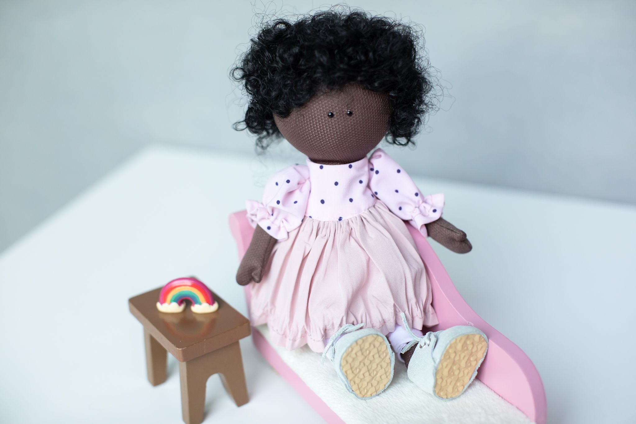 Dark-skinned fabric doll in pink dress sitting on miniature bed with small wooden stool beside her.