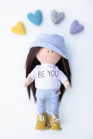 Handmade fabric doll in blue bucket hat and BE YOU sweatshirt surrounded by colorful felt hearts.