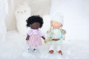 Two handmade fabric dolls, one dark-skinned in pink dress and one fair in mint outfit, in snowy scene.