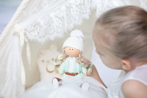 Toddler in white dress examining a handmade mint-colored doll with white hat near a lace bassinet.