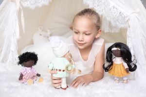 Young girl playing with three diverse handmade cloth dolls inside a white lace teepee tent.