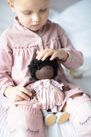 Baby in pink dress gently touching the curly hair of a dark-skinned handmade fabric doll.