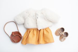 Doll outfit flat lay featuring white fur coat, yellow polka dot skirt, brown bag, and gold bow shoes.