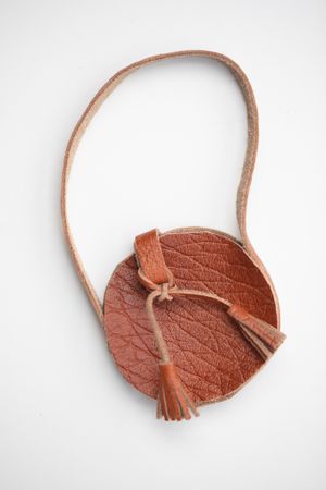 Round brown leather crossbody bag with tassel detail and long strap on white background.